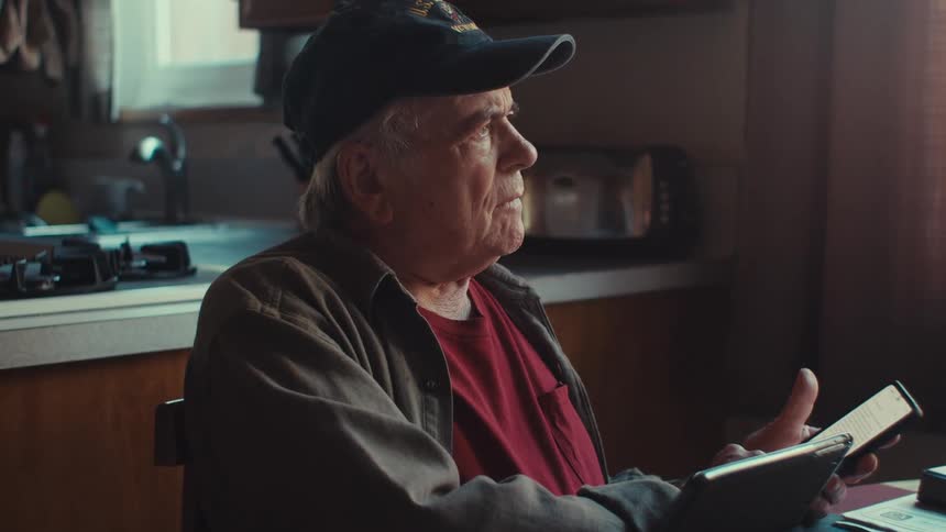 An old man sitting at a dining table by the kitchen types on a smartphone and then stares into the distance