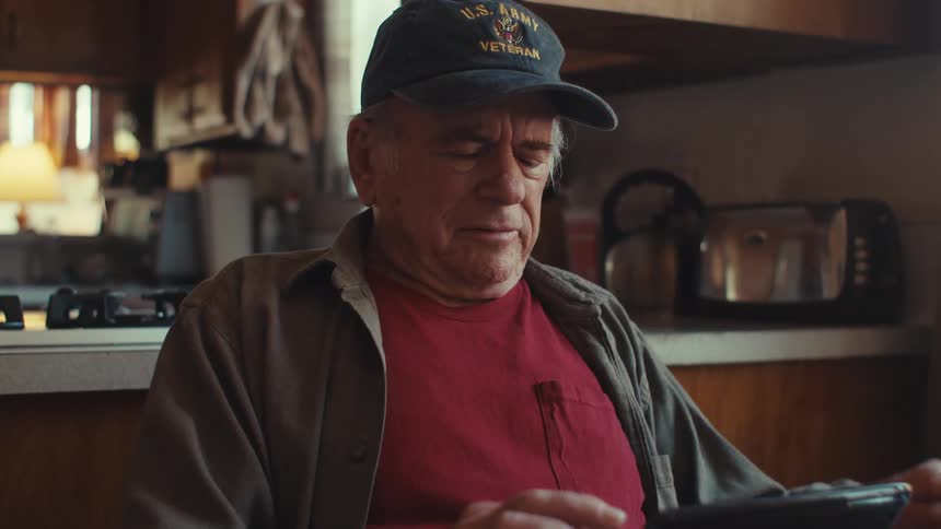 An elderly man wearing a cap sits in a kitchen reading from a tablet