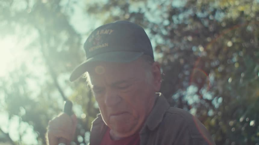 An elderly man standing outdoors in sunlight cleaning with a broom