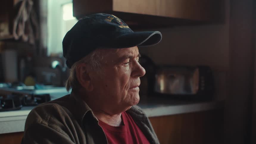 Elderly man frowns while sitting at his kitchen table alone