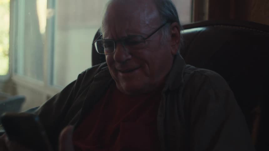 An old man sits indoors laughing at something he sees on a smartphone