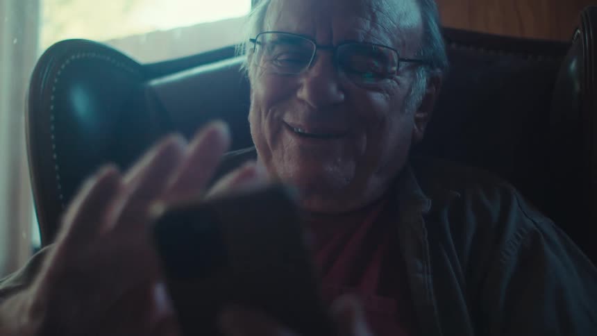 An old man sits in a chair laughing while using a smartphone