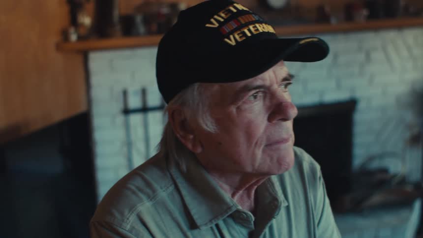 An old man wearing a vietnam veteran hat looks disturbed while standing in front of a fireplace