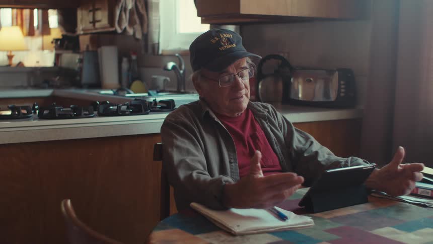Elderly man sitting at his kitchen table reacts while using a tablet for a video call