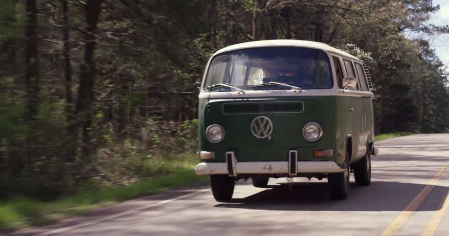 An old vw bus is driving down a road in a forested area
