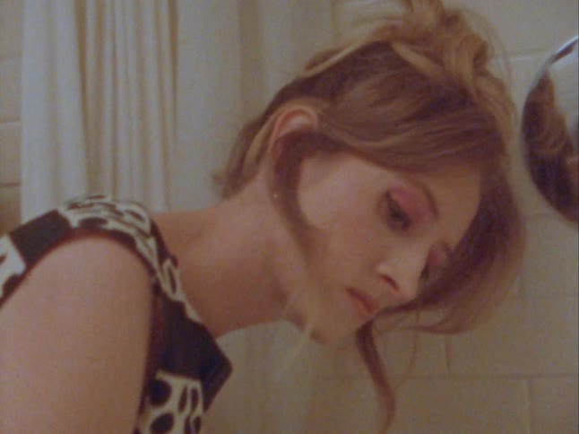 A dirty blonde woman in a polka dot dress looks angry as she scrubs a stain in a bathtub