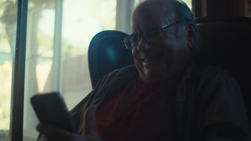 An older man with glasses smiles and waves while looking at his phone sitting in a dimly lit room