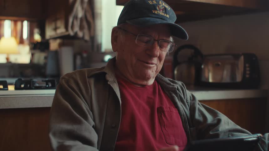 Older man wearing a veteran cap smiles while sitting at home and talking during a video call