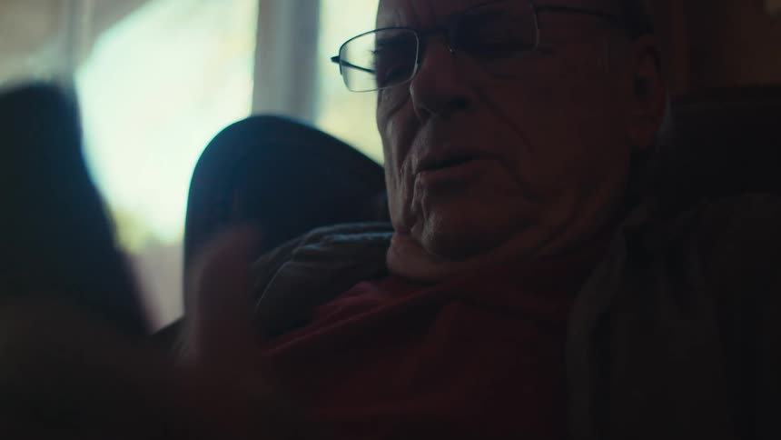 An old man sitting in a chair in a living room looks at a smartphone and starts laughing