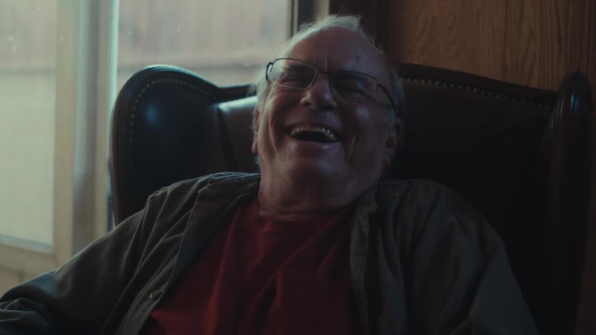 An old man sitting in a padded chair laughs and laughs over something he sees on a smartphone