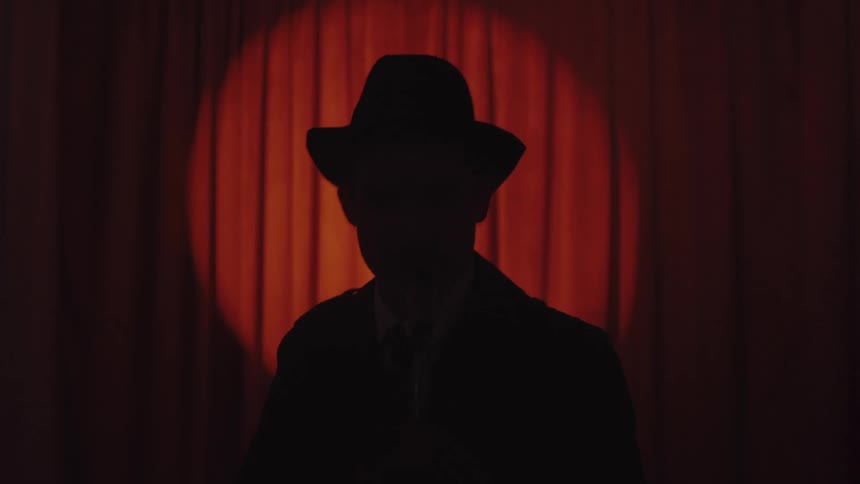 Silhouette of a man in a hat facing a microphone and against a red curtain at a presentation