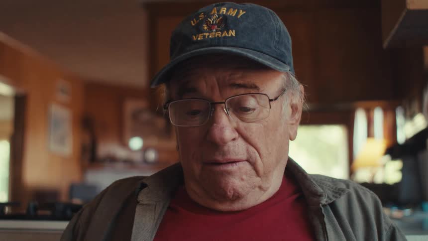 A man wearing glasses and a us army veteran hat sits in a kitchen and speaks