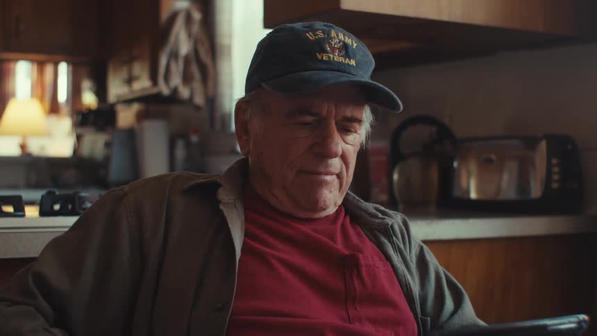 An old man sits in his kitchen reading something on his phone