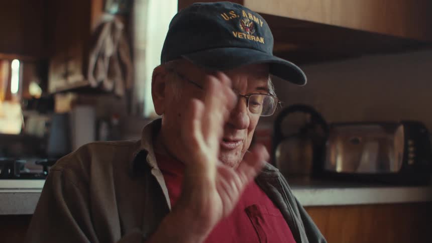 Elderly man smiles and waves while making a video call from the kitchen nook in his home