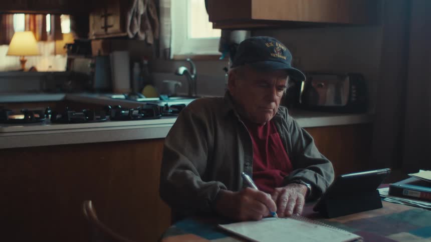 An old man touches a tablet at a dining room table and then begins to write something down from the screen