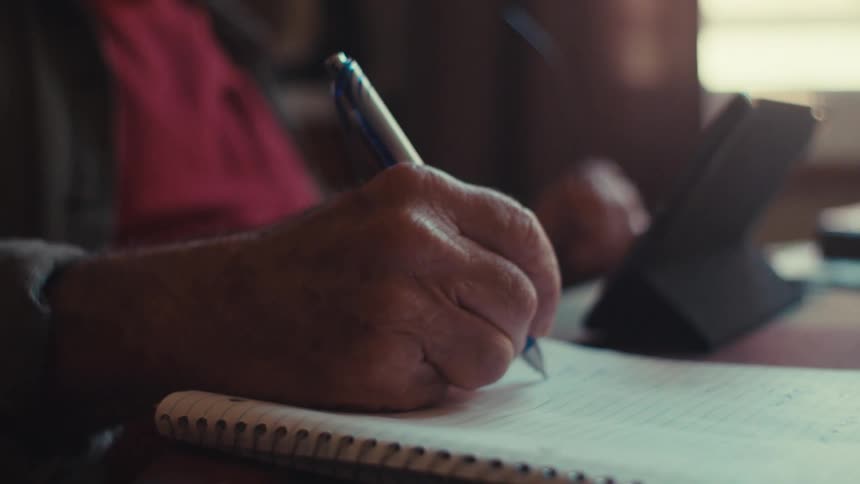 An older man in a us army veteran hat writes in a notebook while looking at a tablet