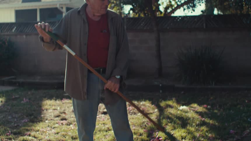 Elderly man standing in a backyard sweeping dry fallen leaves from the grass