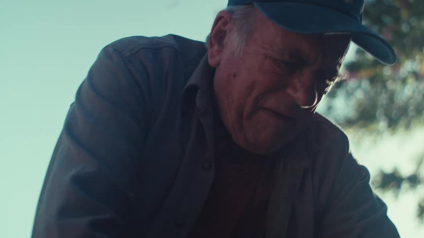 An old man uses a rake to lift leaves off of a yard