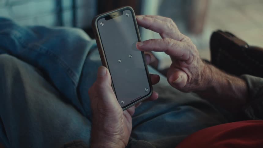 An older man is sitting indoors while scrolling on his smartphone
