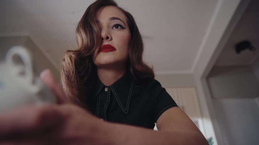 A woman with 1940s style hair and makeup is shown closely examining a small white cup