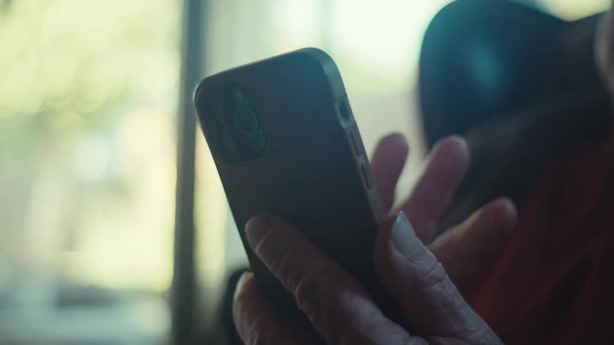 An elderly man sits indoors while scrolling on a smartphone
