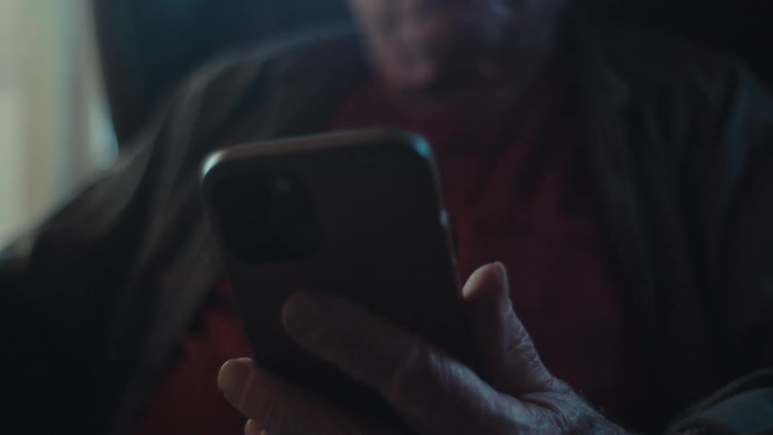 An old man sits in a padded chair in a living room and talks to a smartphone