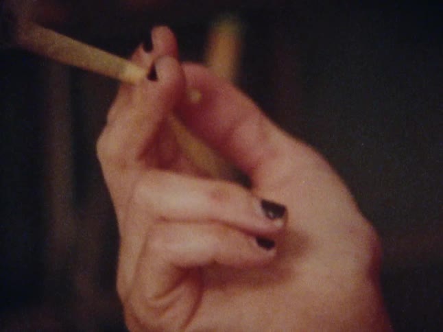 A redhaired woman in a red shirt is using a candle to light a cigarette and smoke it
