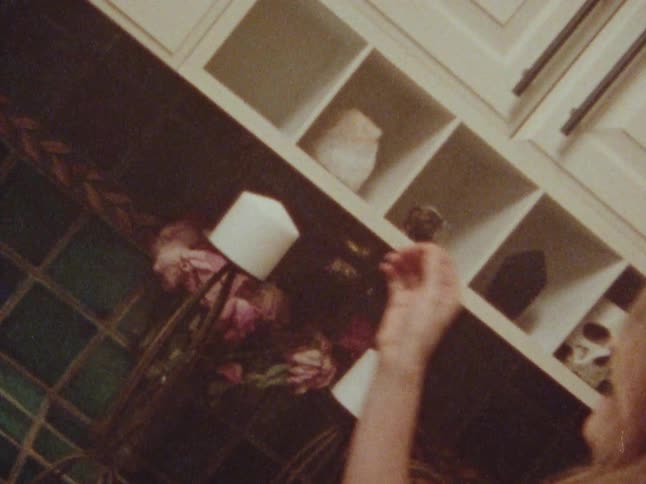 A woman with long blonde hair lights a candle in a kitchen with a skull and other items on a shelf in the background