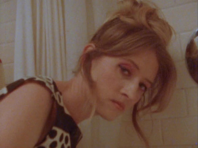 A woman in a dress scrubs water stains in a tub while looking fearful