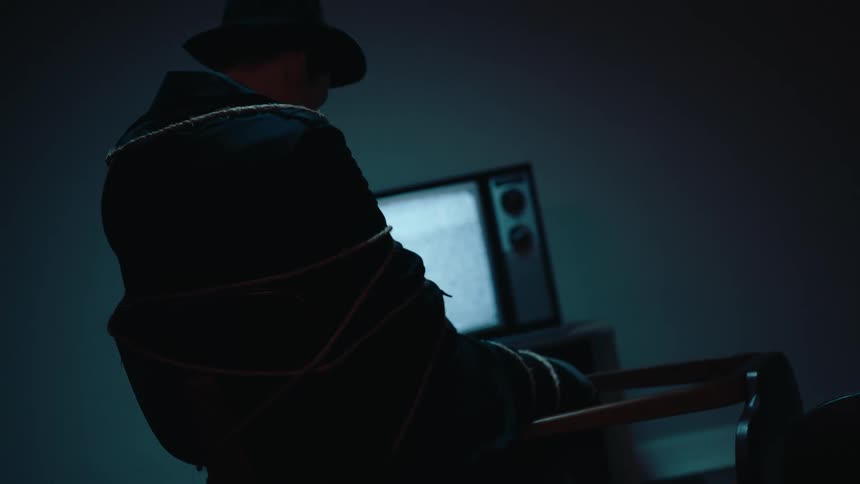 Man tied to a chair in a dark room with a television in the background with no signal