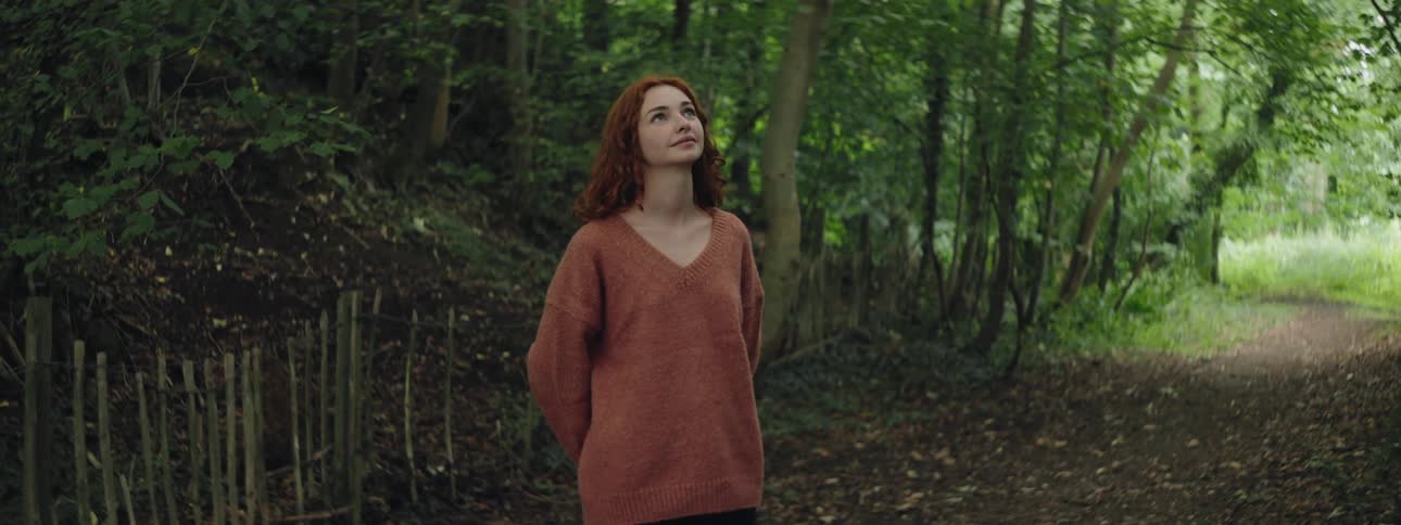 A young woman with red hair and red clothes is looking up at an area where there is a dirt path and trees behind