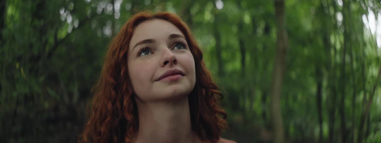 The face of a young redhaired woman who is looking up and behind her there are several green leafy trees