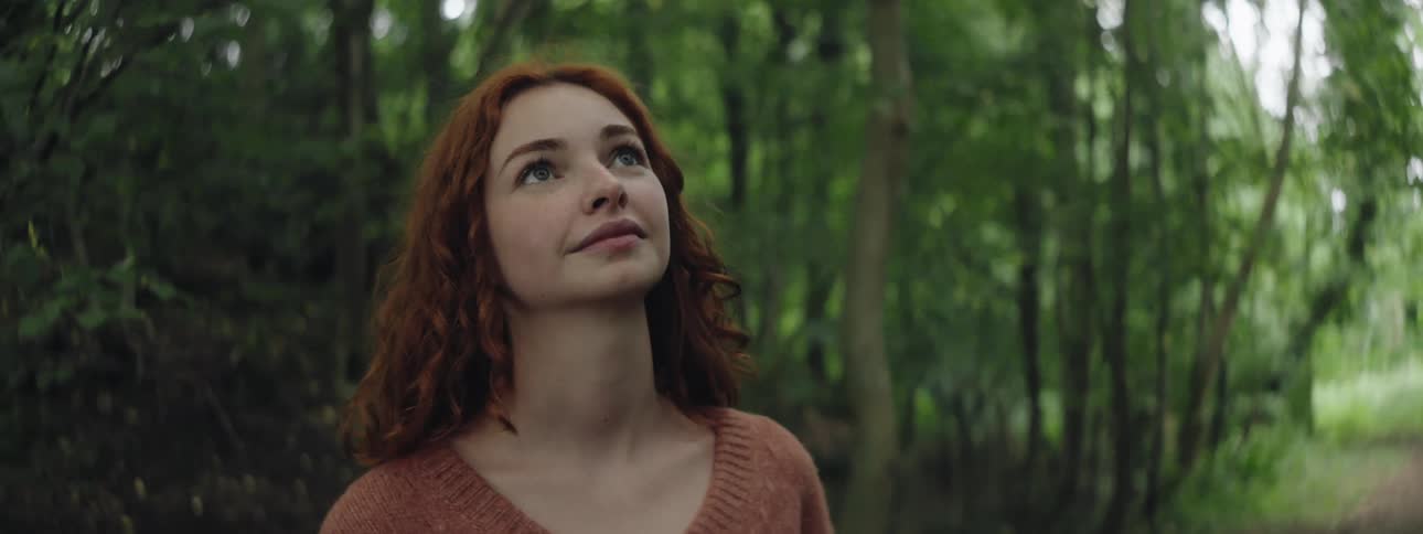 A happy woman with red hair and brown sweater looks up in the forest