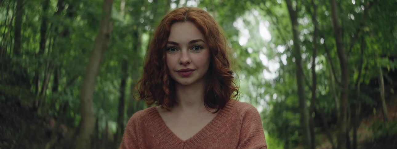 A woman with red curly hair stands in the woods wearing a brown sweater
