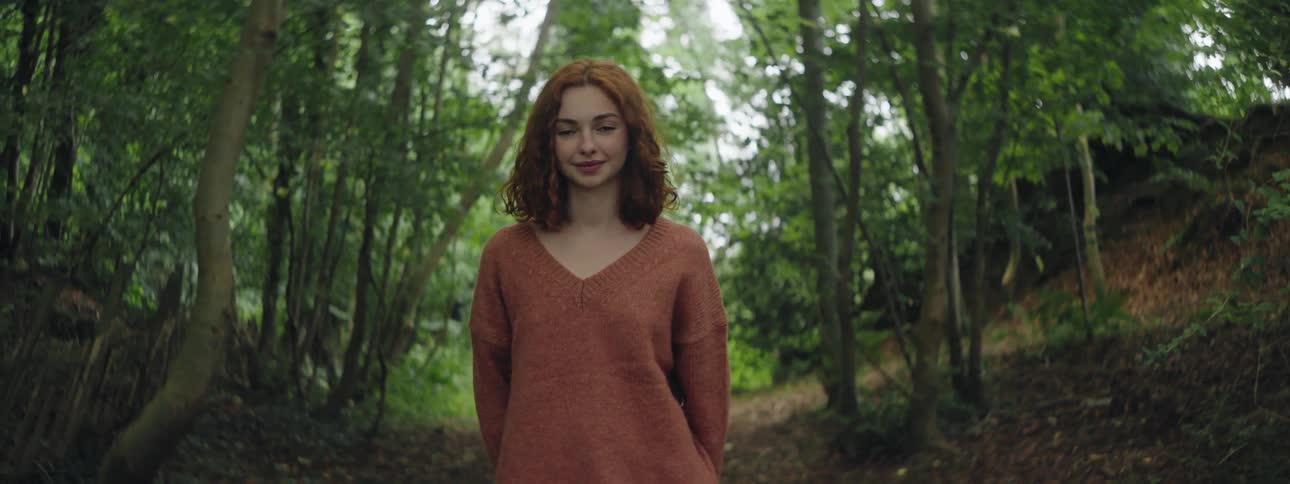 A teenage girl with red hair and a sweater smiles as she stands on a path in a forest