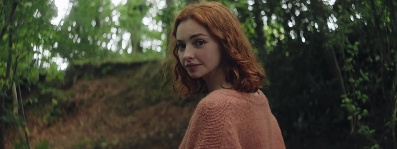 A young woman with wavy hair looks over her shoulder while standing in a grove of trees