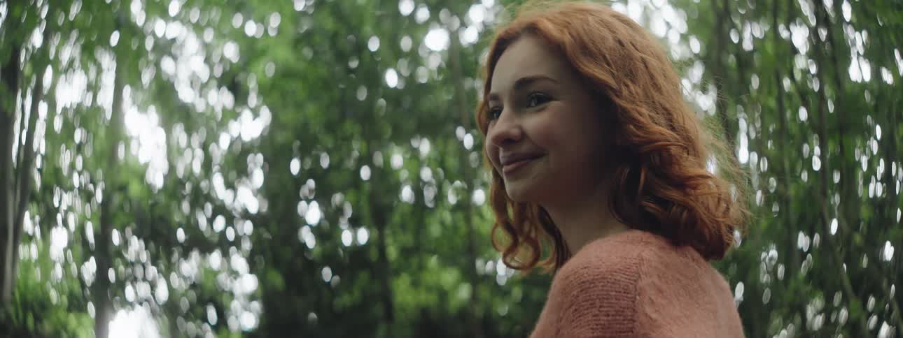A redhead teen smiles and blinks while standing in a forest