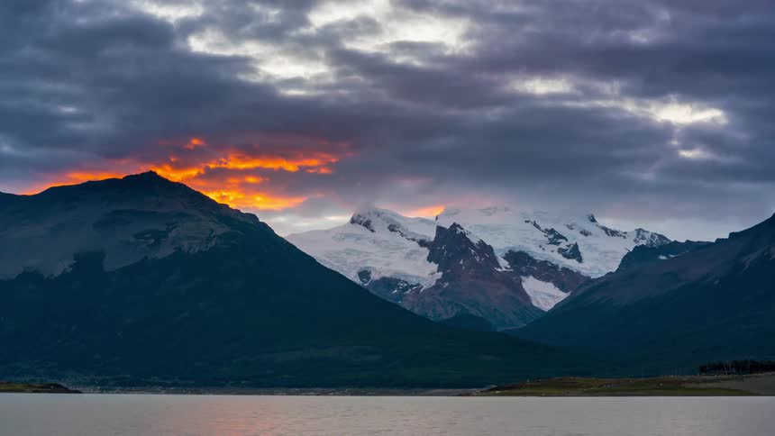 The sun shines behind dark storm clouds floating over a mountainous coastal area
