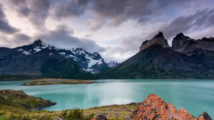 Storm clouds move over a large mountain range next to a light blue lake