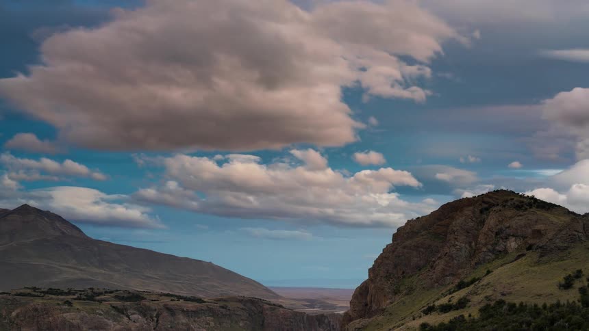 A dramatic landscape with rugged mountains framing a valley under a sky filled with fluffy clouds against a deep blue background