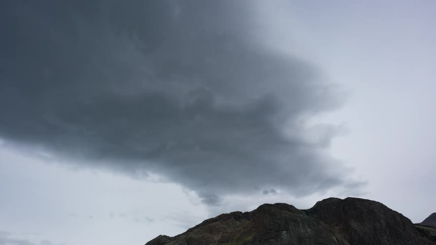 Gray clouds passing quickly in the sky that is whitish and below there is the peak of a rocky mountain