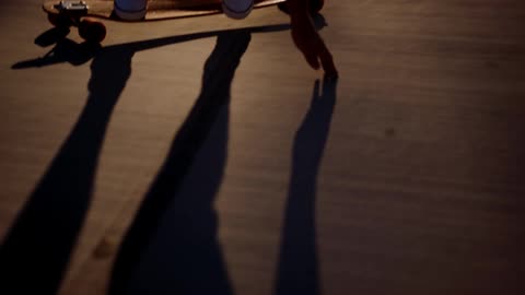 A long shadow of a person slowly rides by the camera on their long skateboard at sunset while touching the ground