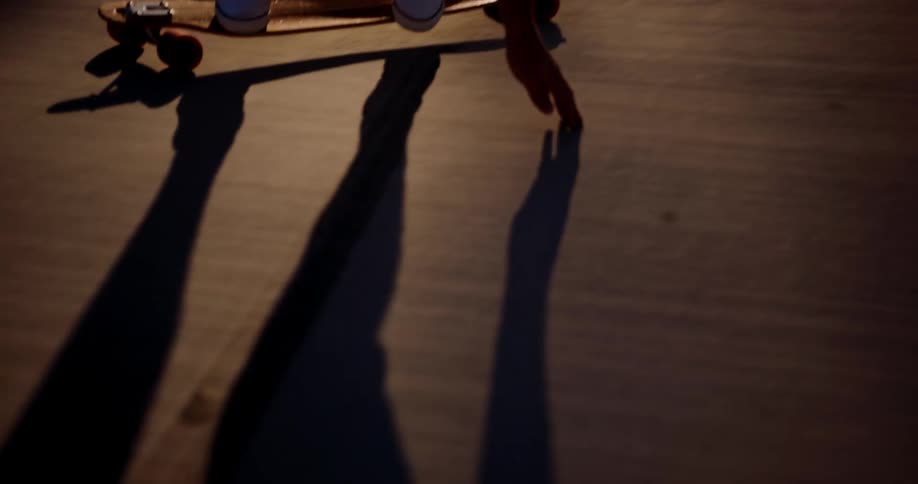 A long shadow of a person slowly rides by the camera on their long skateboard at sunset while touching the ground