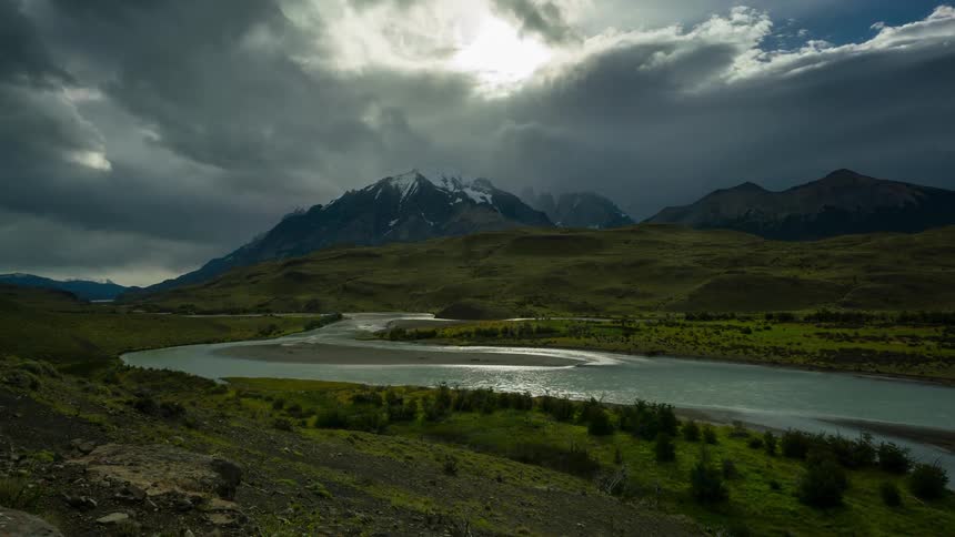 A dramatic landscape in patagonia with a winding river snaking through a lush green valley framed by snowcapped mountains and a dramatic stormy sky