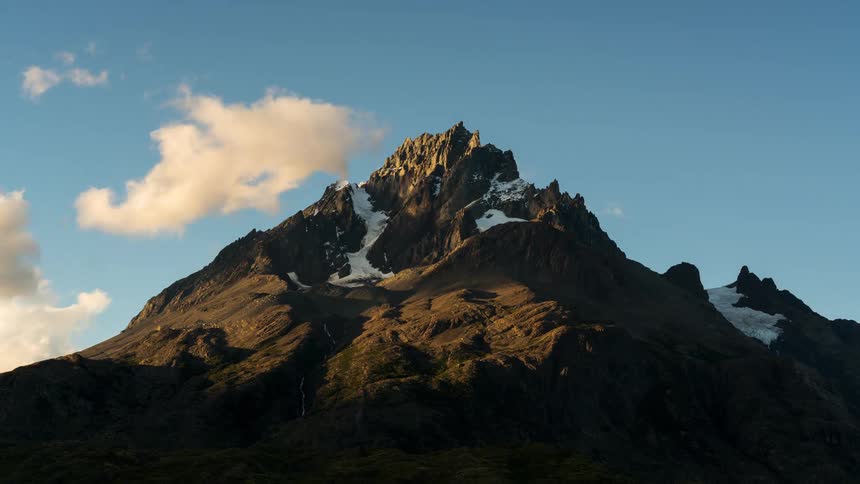 A big rocky mountain with some snow on its top and there are many white clouds passing quickly in the blue sky