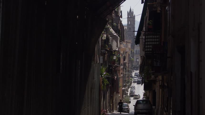 Traffic drives down a narrow city street near buildings and a church