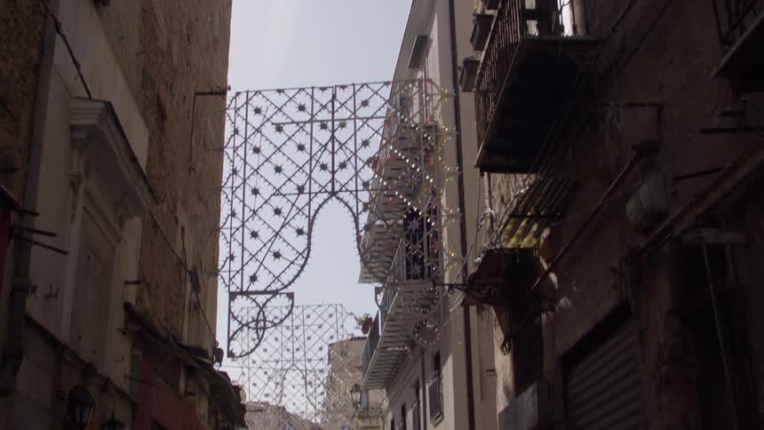 The facade of some buildings with balconies on an alley with some parked cars and two people walking in front