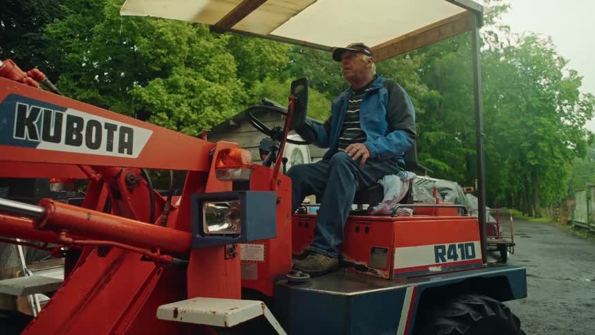 An elderly man wearing a cap is sitting on an orange tractor and looking forward while checking the roof of the tractor
