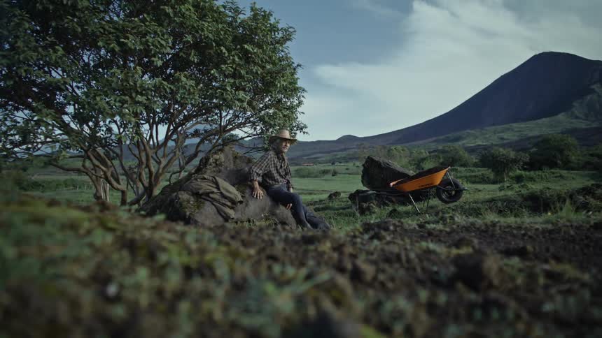 A man is resting on a rock by a wheelbarrow
