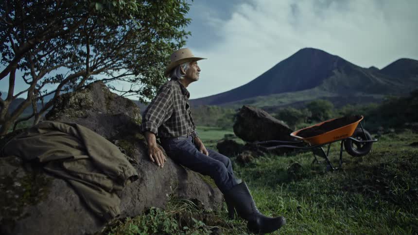 An old man in a hat is sitting on a rock and there is an orange wheelbarrow on the grass in front of the man
